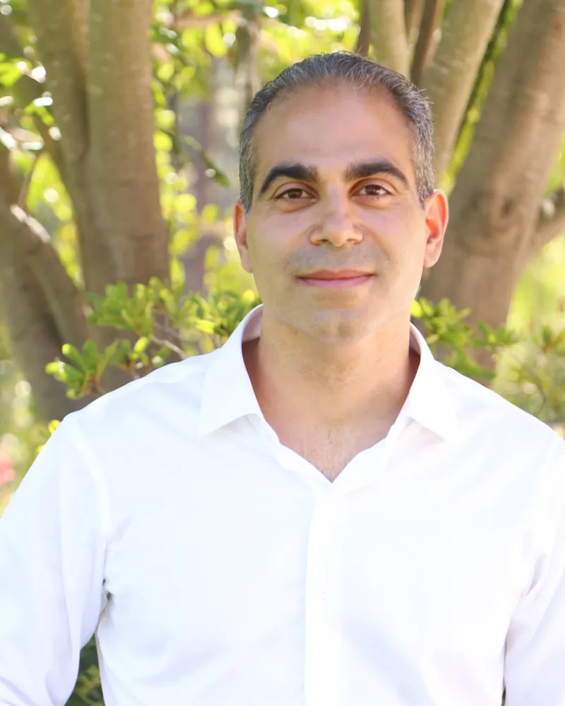 Allen Kamrava, author of The Measure, outdoors in a white shirt in front of a sunlit tree.